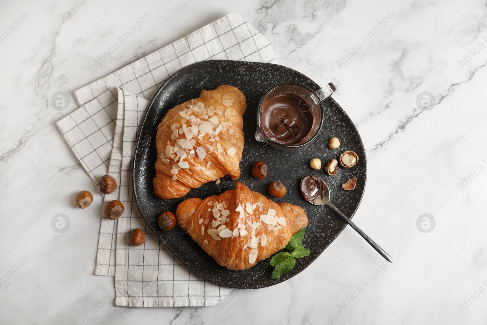 Photo of Delicious croissants with chocolate, nuts and spoon on white marble table, flat lay