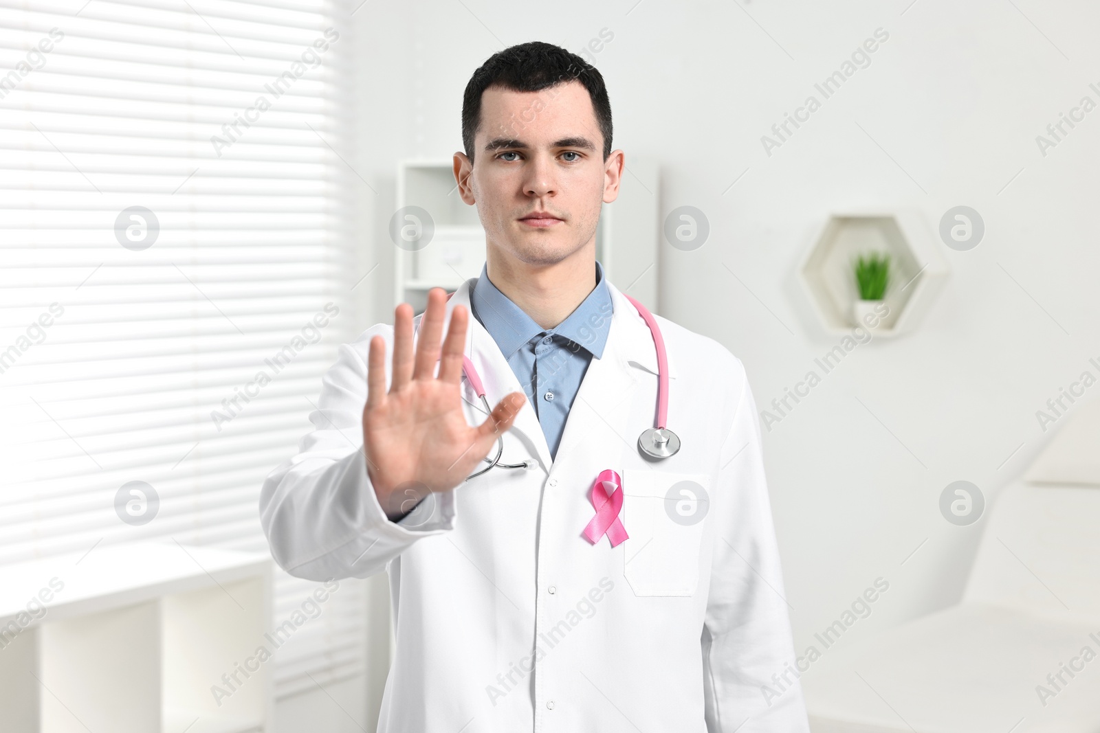 Photo of Mammologist with pink ribbon and stethoscope showing stop gesture in hospital. Breast cancer awareness