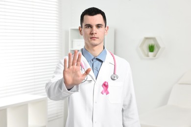 Mammologist with pink ribbon and stethoscope showing stop gesture in hospital. Breast cancer awareness