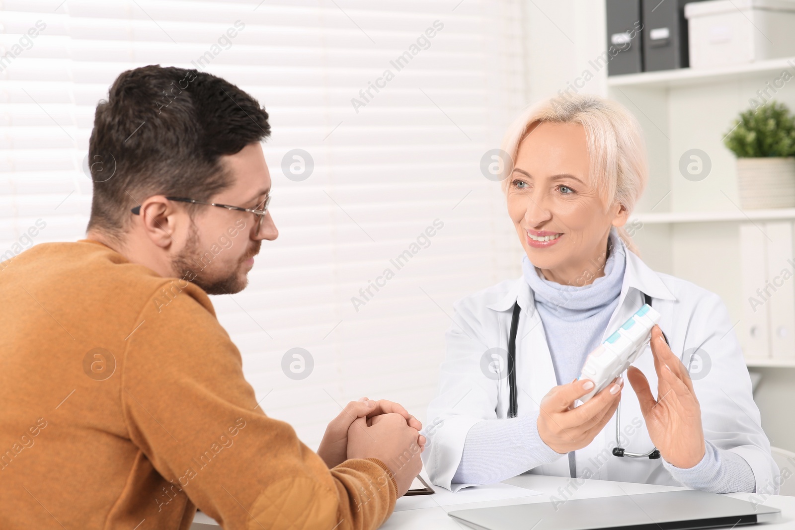 Photo of Doctor holding box of pills and consulting patient at white table in clinic