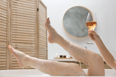 Woman with glass of wine taking bath in tub indoors, closeup