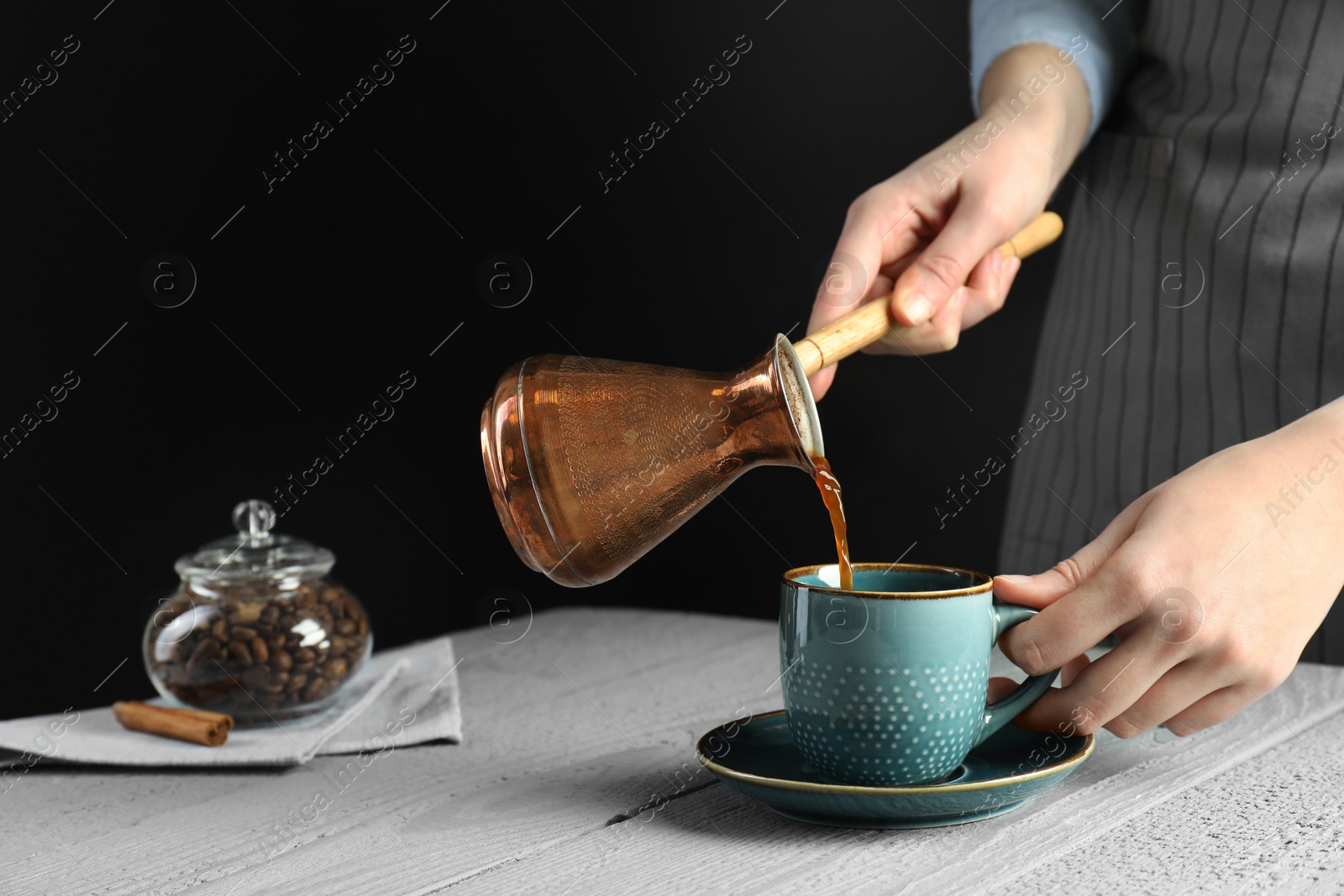 Photo of Turkish coffee. Woman pouring brewed beverage from cezve into cup at gray wooden table against black background, closeup