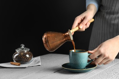 Photo of Turkish coffee. Woman pouring brewed beverage from cezve into cup at gray wooden table against black background, closeup