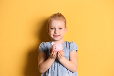 Little girl with piggy bank on color background