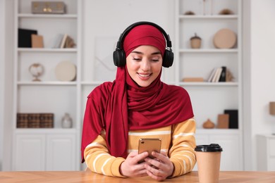 Muslim woman in hijab using smartphone at wooden table in room