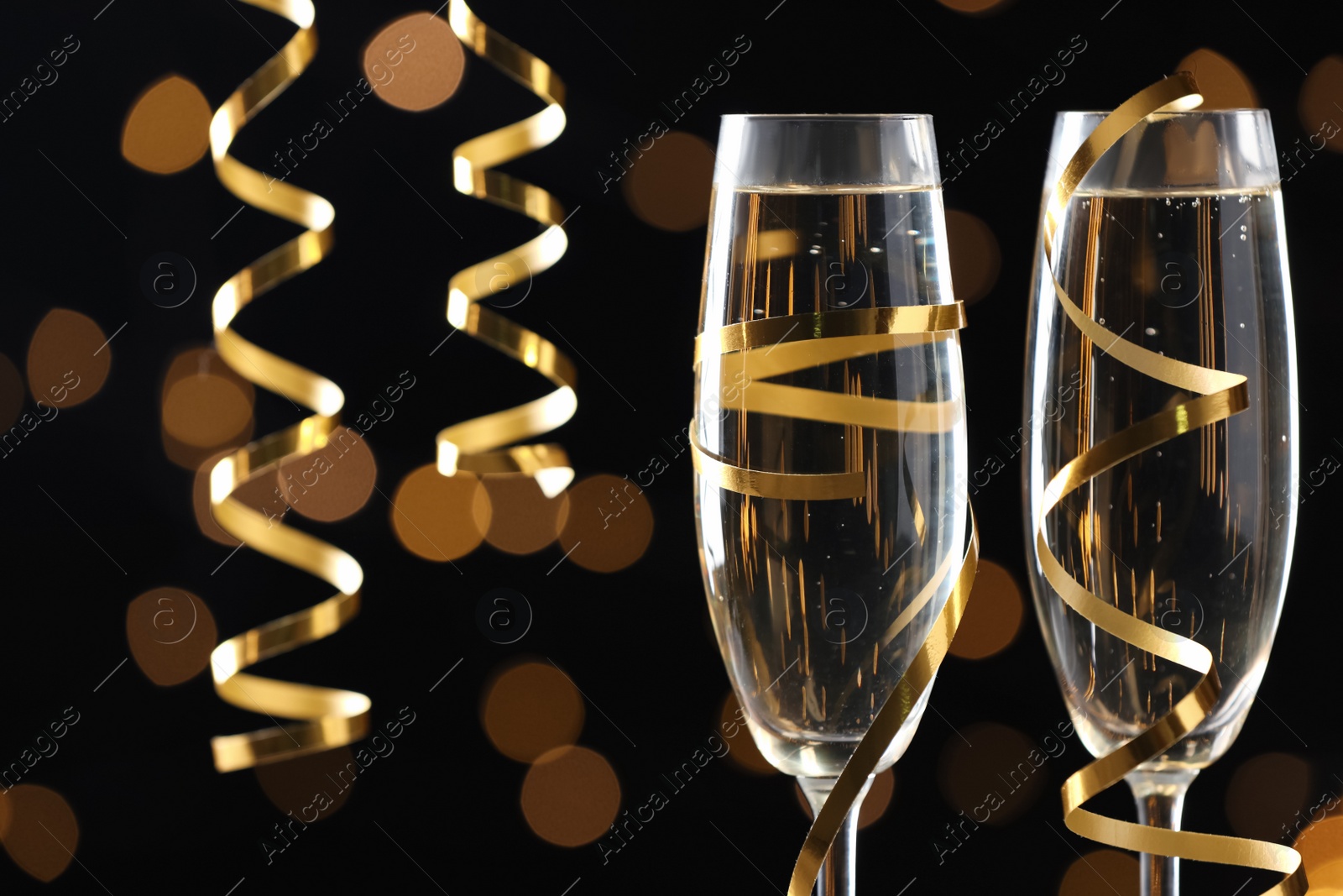 Photo of Glasses of champagne and serpentine streamers against black background with blurred lights, closeup. Space for text