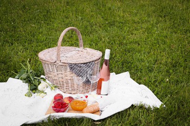 Picnic blanket with tasty food, flowers, basket and cider on green grass outdoors