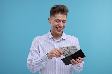 Happy man putting dollar banknotes into wallet on light blue background