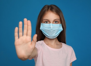 Photo of Little girl in protective mask showing stop gesture on blue background. Prevent spreading of coronavirus