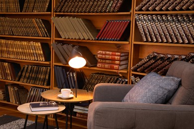 Photo of Book with glasses, table lamp and cup of drink near comfortable armchair in cozy home library