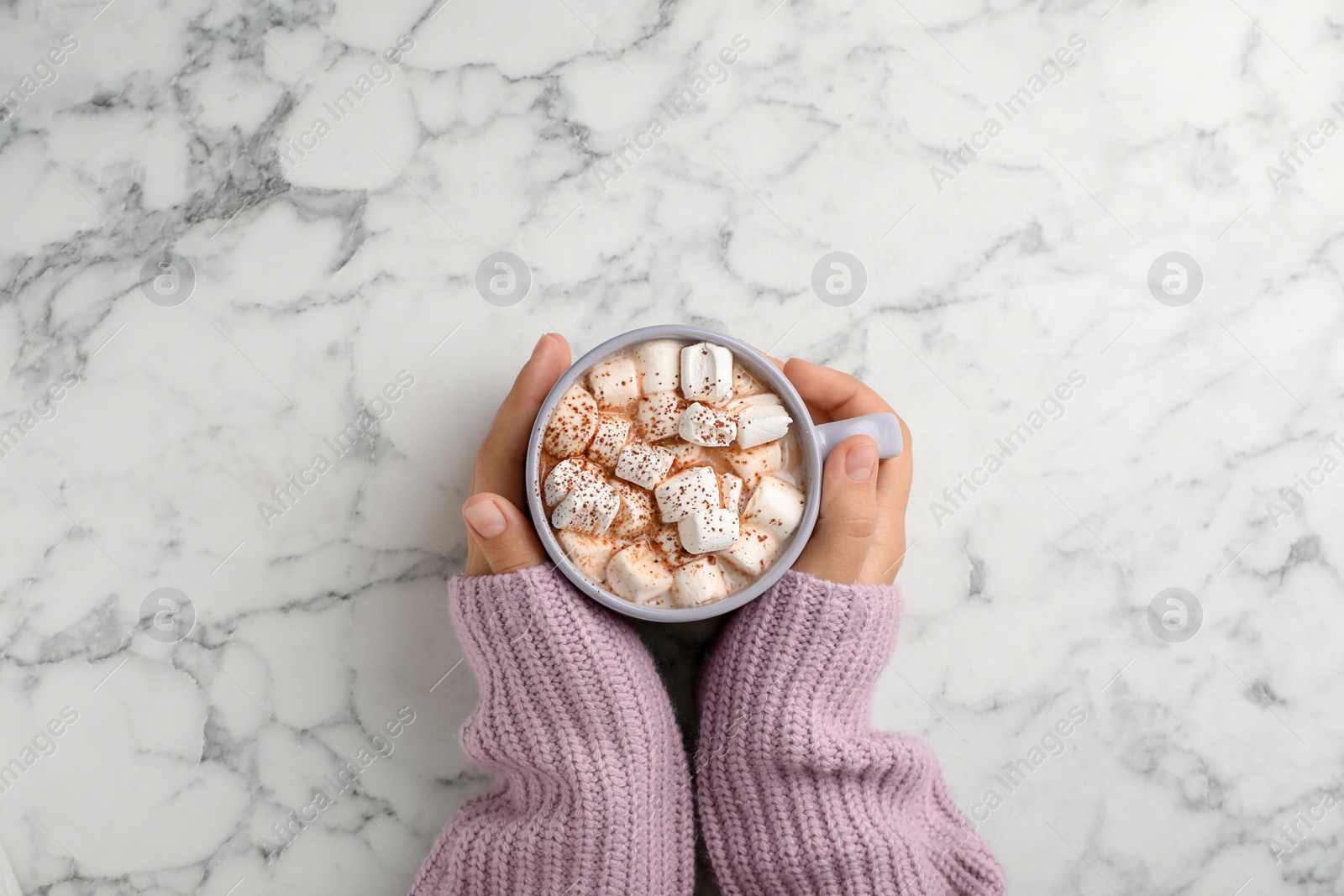 Photo of Woman in sweater holding cup of tasty chocolate with milk and marshmallows on marble table, top view
