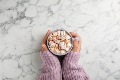 Photo of Woman in sweater holding cup of tasty chocolate with milk and marshmallows on marble table, top view