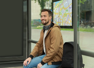Image of Young man with headphones and backpack waiting for public transport at bus stop