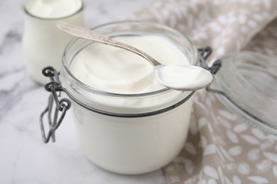 Delicious natural yogurt in glass jar and spoon on white marble table, closeup