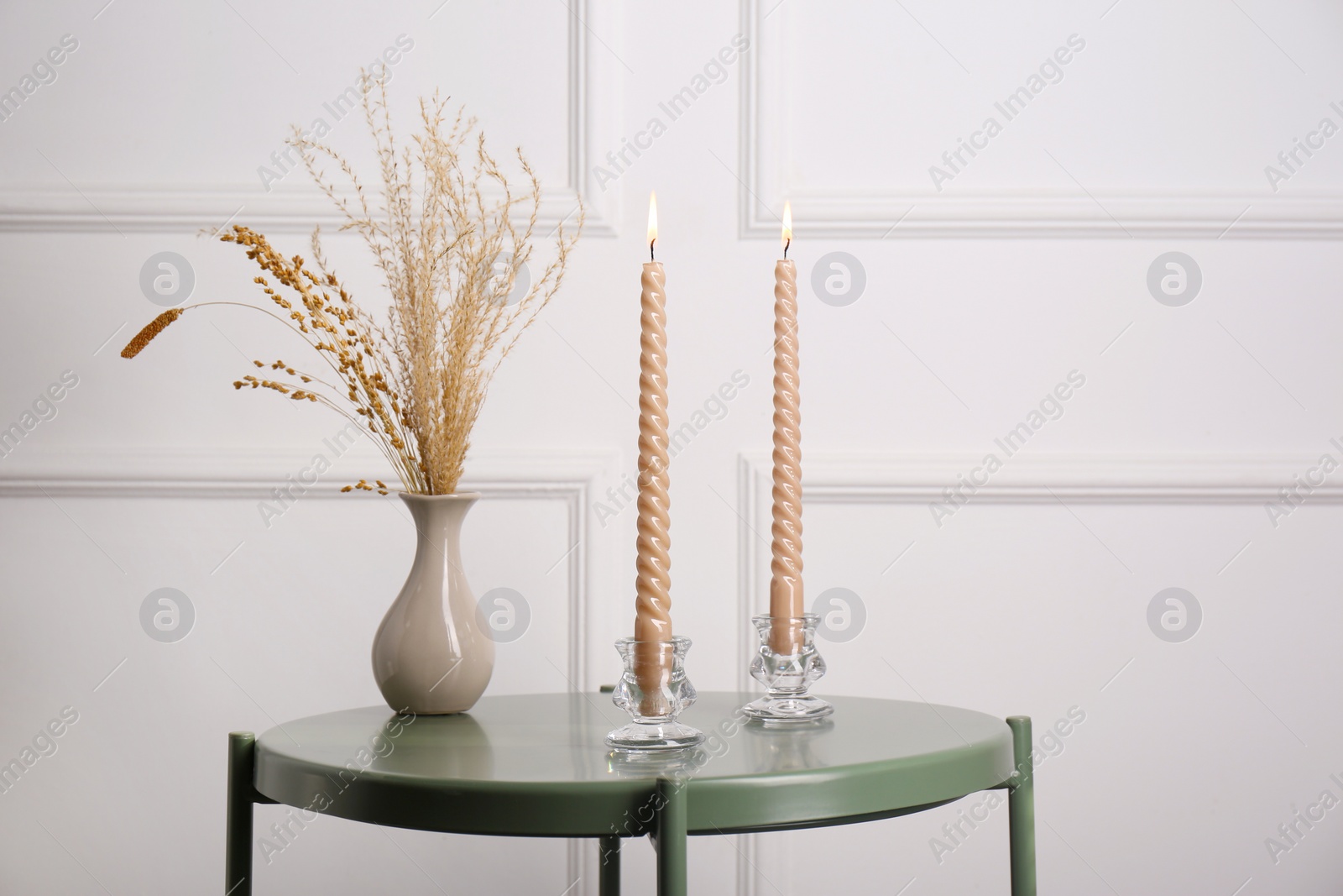 Photo of Dry plants in vase and burning candles on table near white wall