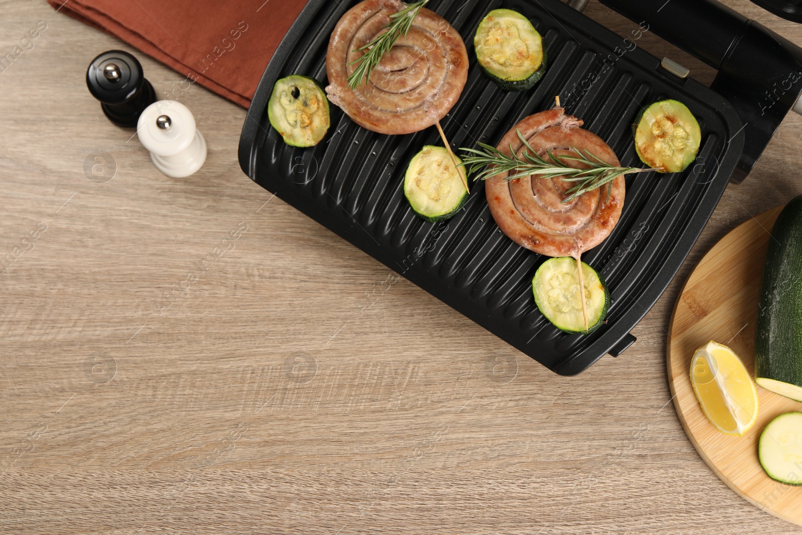 Photo of Electric grill with homemade sausages, zucchini and rosemary on wooden table, flat lay. Space for text