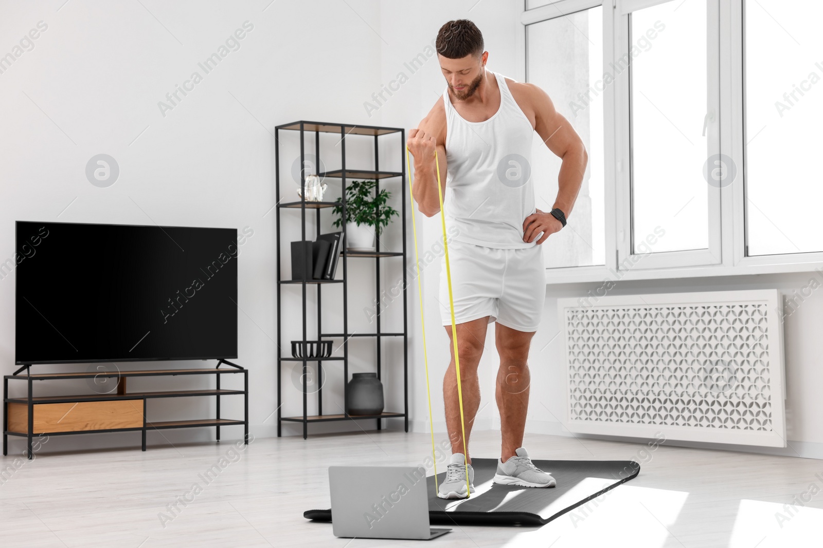 Photo of Muscular man doing exercise with elastic resistance band near laptop on mat at home