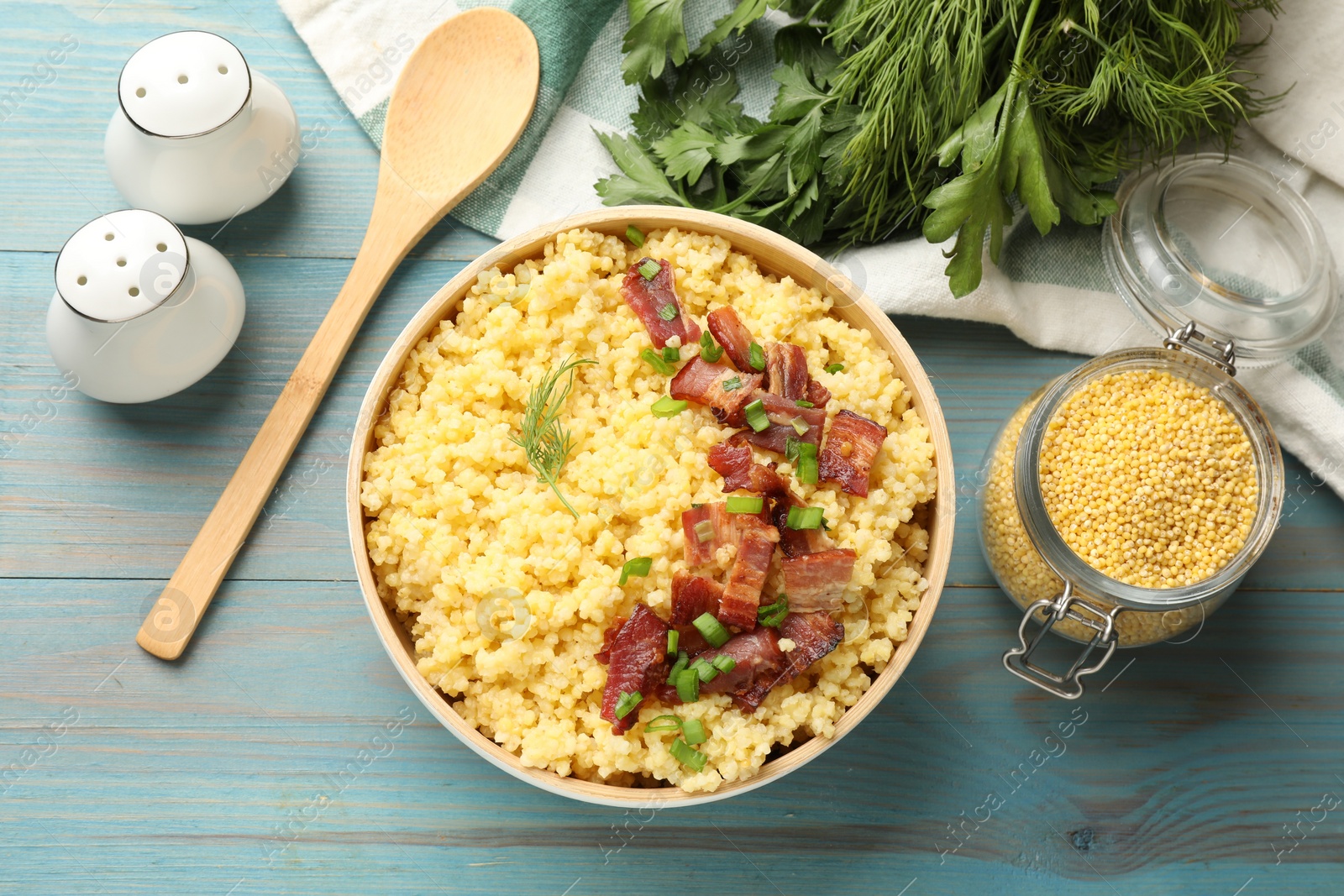 Photo of Tasty millet porridge with bacon, green onion and dill in bowl served on light blue wooden table, flat lay