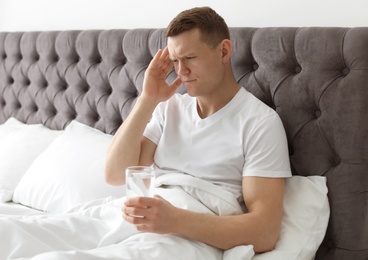 Photo of Young man with terrible headache holding pills and glass of water in bed