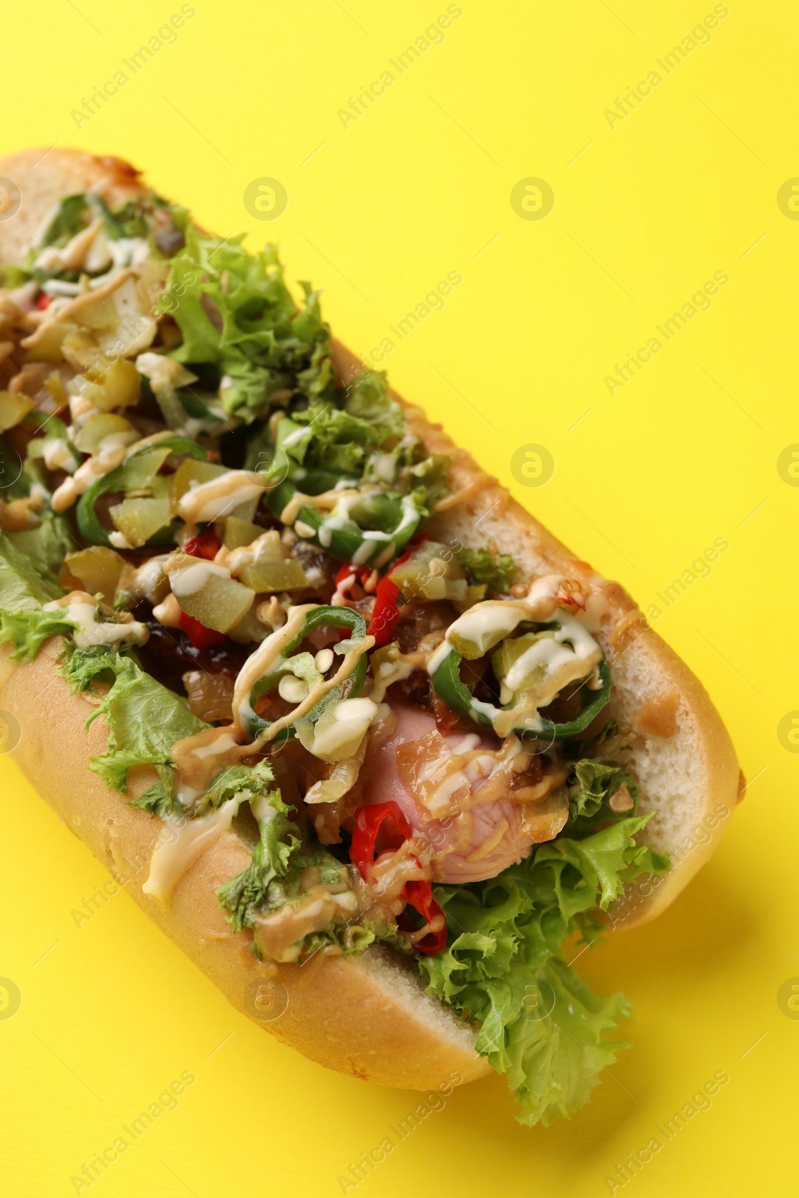 Photo of Delicious hot dog with chili peppers, lettuce, pickles and sauces on yellow background, closeup