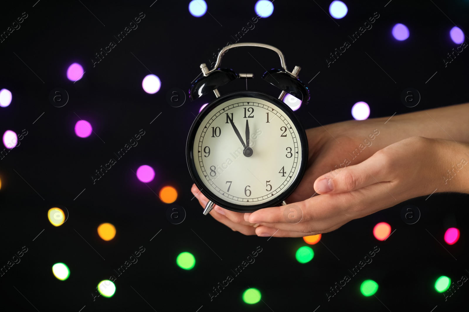 Photo of Woman holding alarm clock against blurred lights, closeup with space for text. New Year countdown