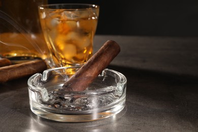 Photo of Smoldering cigar, ashtray and whiskey on grey table, closeup. Space for text