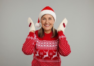 Happy senior woman in Christmas sweater, Santa hat and knitted mittens on grey background