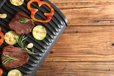 Photo of Electric grill with tasty meat steaks, rosemary and vegetables on wooden table, top view. Space for text