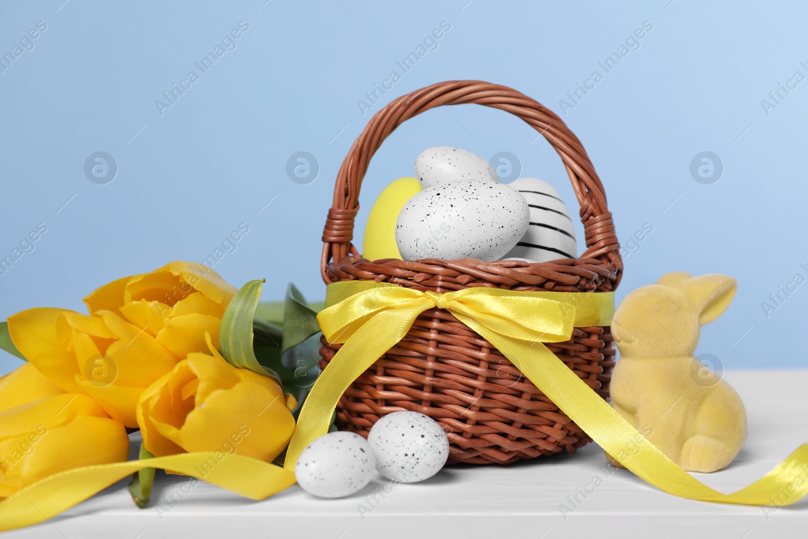 Photo of Wicker basket with festively decorated Easter eggs, bunny and beautiful tulips on white wooden table against light blue background