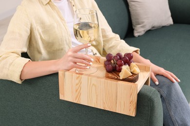 Woman holding glass of wine and snacks on sofa armrest wooden table at home, closeup