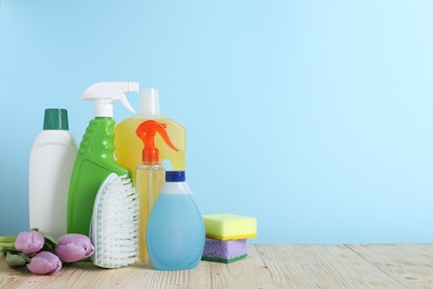 Photo of Spring cleaning. Different detergents, tools and beautiful flowers on wooden table against light blue background, space for text