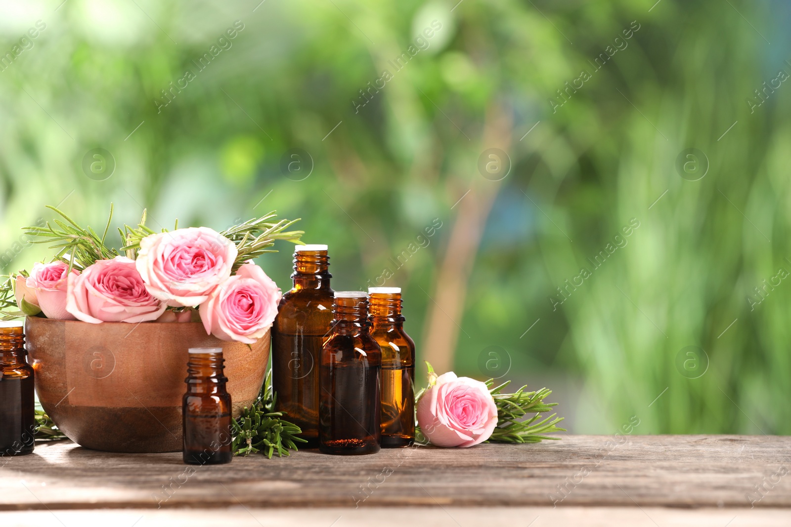 Photo of Bottles with essential oils, roses and rosemary on wooden table against blurred green background. Space for text