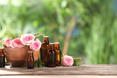 Bottles with essential oils, roses and rosemary on wooden table against blurred green background. Space for text