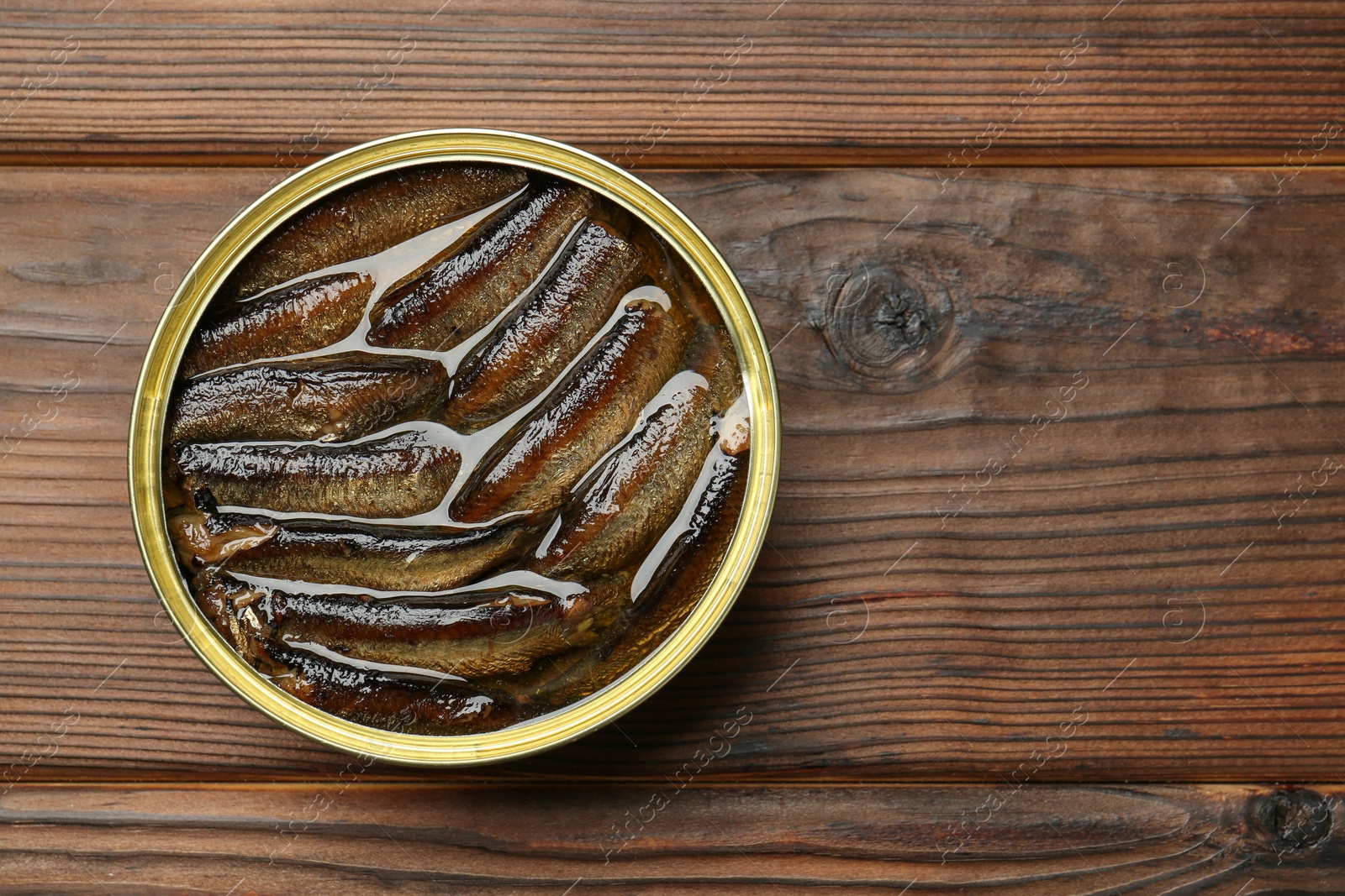 Photo of Open tin can of sprats on wooden table, top view. Space for text