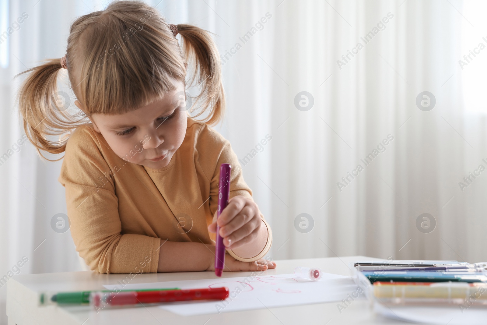 Photo of Cute little girl drawing with marker at white table indoors. Child`s art