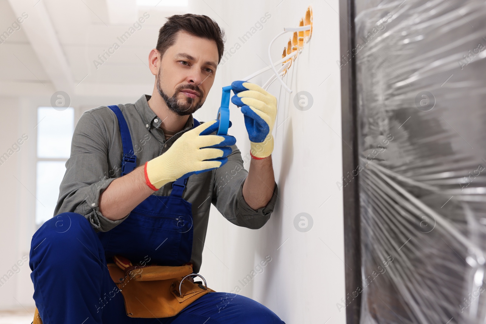 Photo of Electrician in uniform with insulating tape repairing power socket indoors