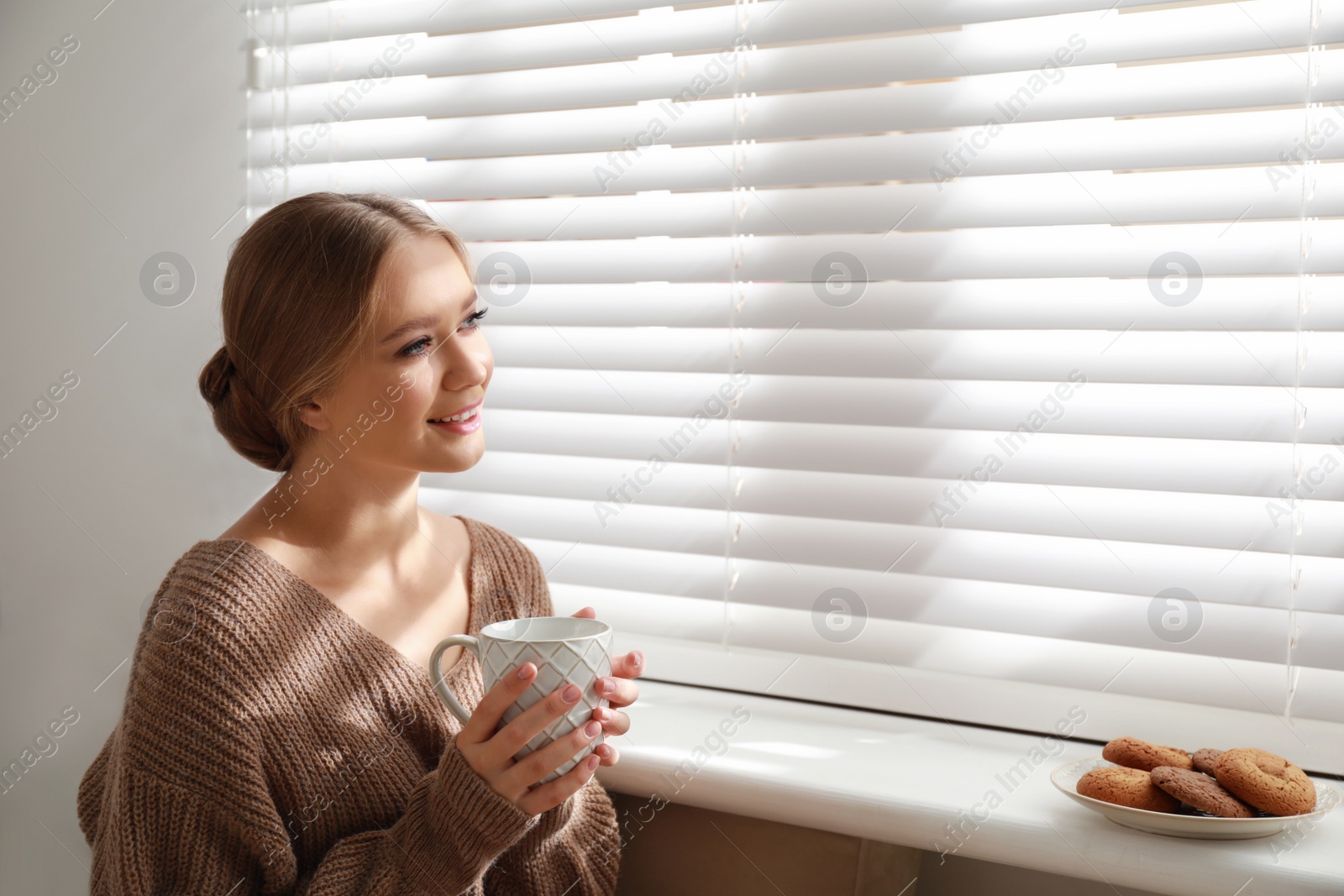 Photo of Beautiful young woman with cup of hot drink and cookies near window at home. Winter atmosphere