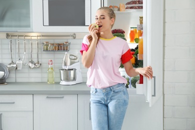 Woman with apple standing near open refrigerator in kitchen