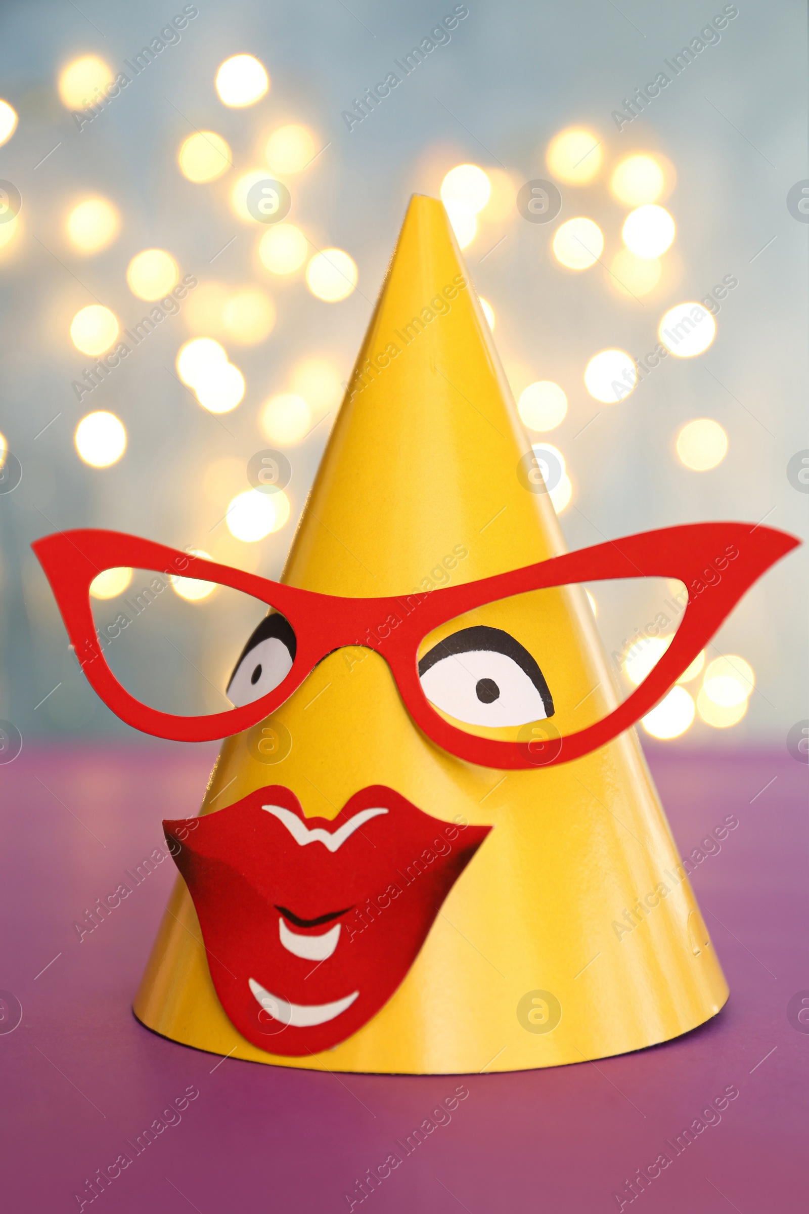 Photo of Funny handmade party hat with glasses and lips on purple table against blurred lights, closeup