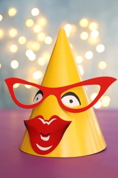 Funny handmade party hat with glasses and lips on purple table against blurred lights, closeup