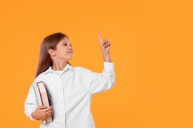 Cute schoolgirl with books pointing at something on orange background. Space for text