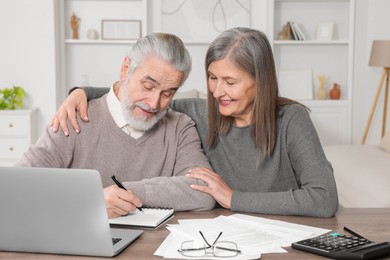 Elderly couple with papers and laptop discussing pension plan at wooden table in room