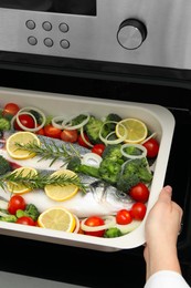 Photo of Woman putting baking dish with raw fish and vegetables into oven, closeup