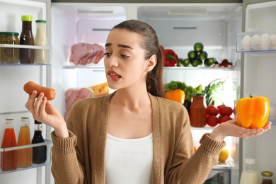 Photo of Young woman with sausage and bell pepper near open refrigerator