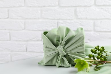 Furoshiki technique. Gift packed in green fabric and plants for decor on white table, space for text