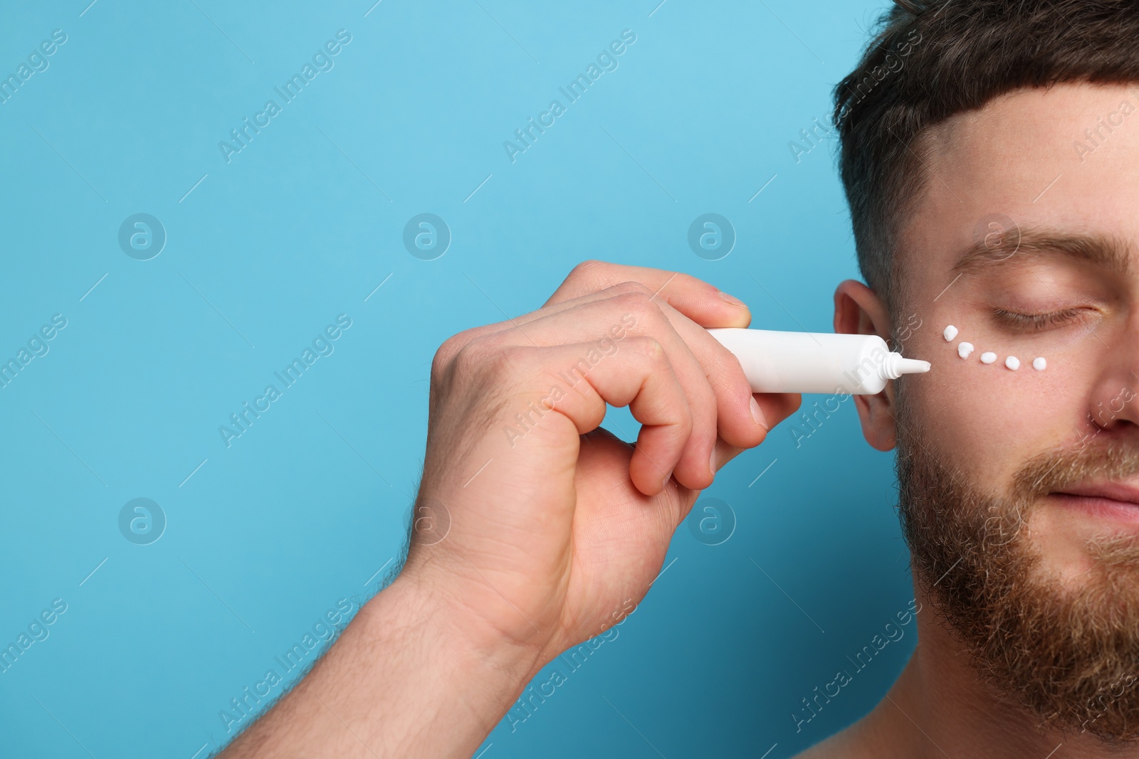 Photo of Handsome man applying moisturizing cream onto his face on light blue background, closeup