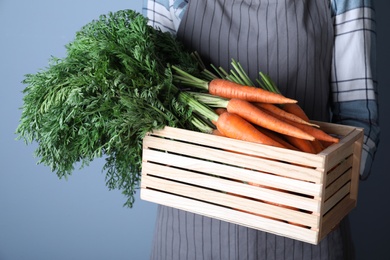 Photo of Woman holding wooden crate with ripe carrots on light blue background, closeup