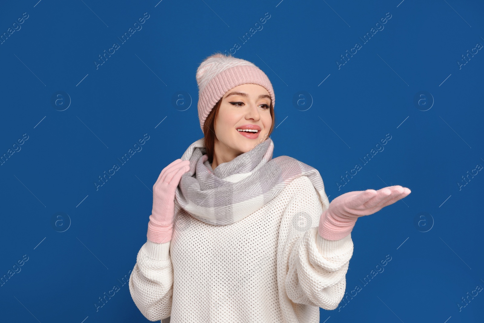 Photo of Young woman wearing warm sweater, gloves, scarf and hat on blue background. Winter season