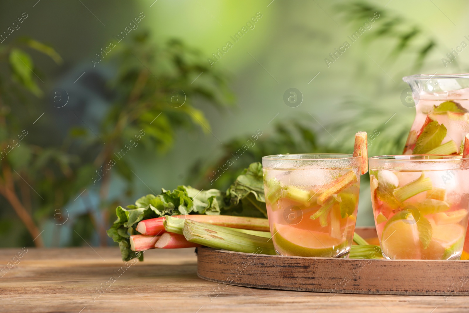 Photo of Glasses and jug of tasty rhubarb cocktail with citrus fruits on wooden table outdoors, space for text