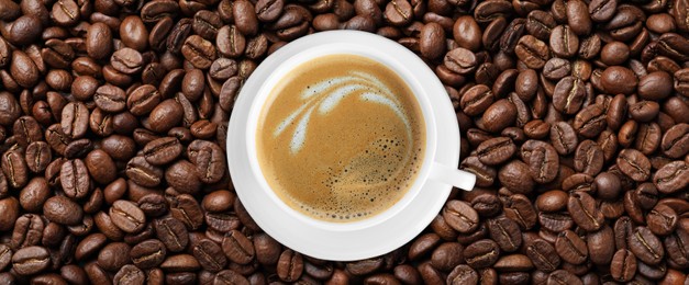 Image of Cup of tasty aromatic coffee on roasted beans, top view. Banner design